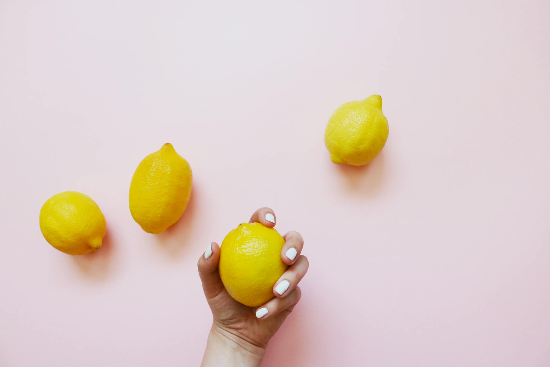 A hand holding a fresh yellow lemon on a soft pink background with additional lemons nearby, symbolizing freshness and sensitivity to touch.