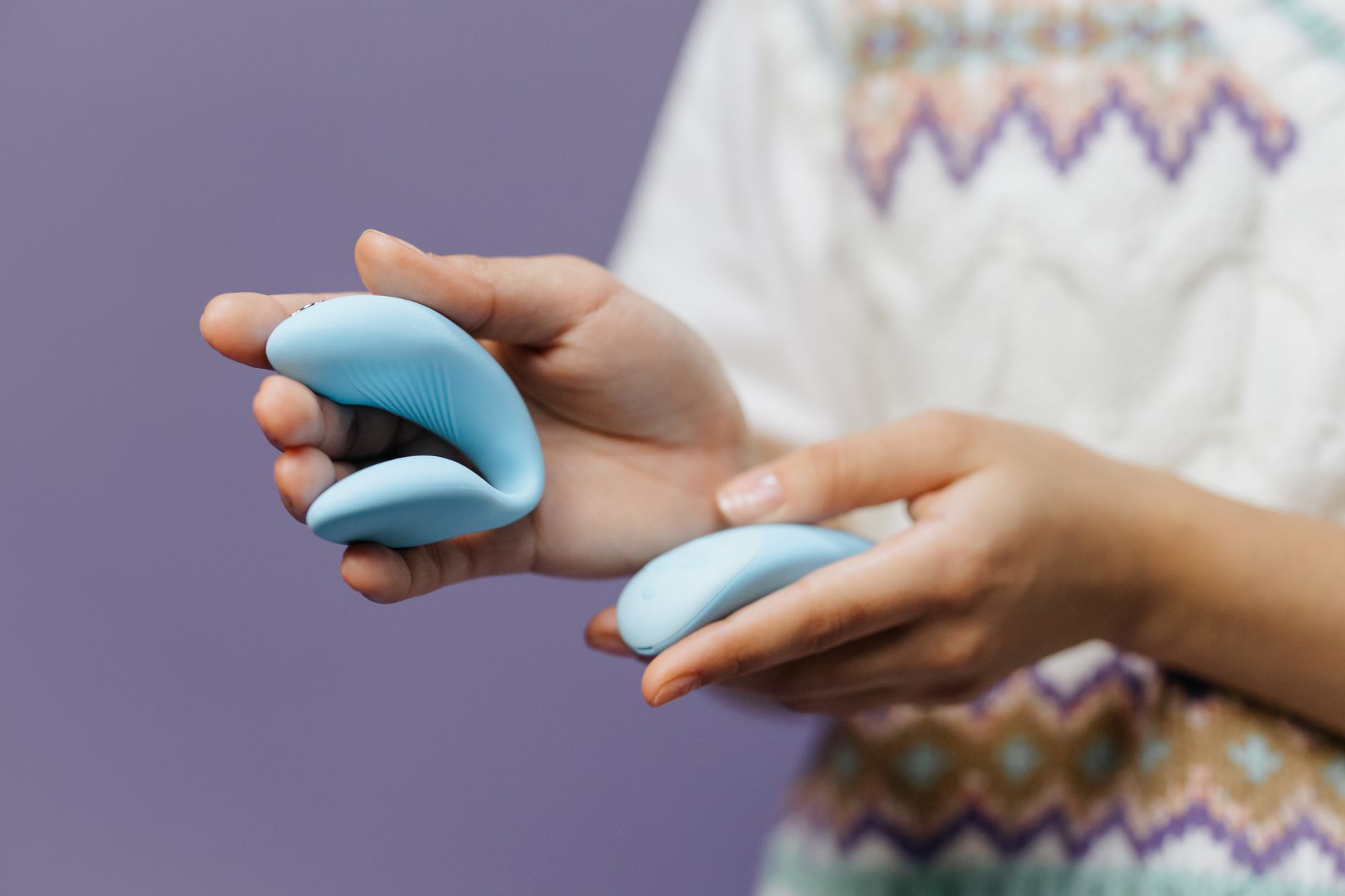 Close-up hands holding a sleek blue clitoral vibrator against a purple background